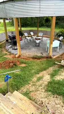 Outdoor covered patio under construction with wooden posts, corrugated metal walls, gravel ground, white folding chairs, grill, and mint-green cabinet.