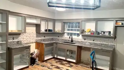 In-progress kitchen with gray cabinets, granite countertops, white subway tile backsplash, and a sink under a window.