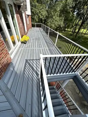 Long gray deck with white railings along brick house; yellow flowers by sliding door; green lawn and trees.