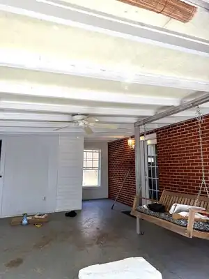 Brick-walled enclosed porch with white ceiling beams, ceiling fan, and a wooden porch swing.